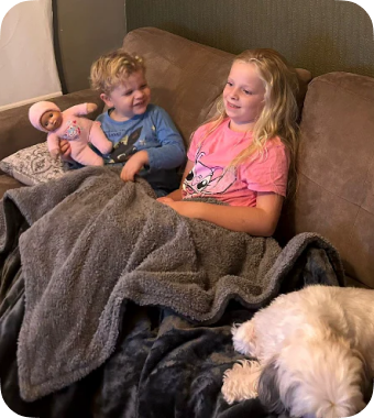 Twee kinderen en een hond liggen samen onder een warme Seranova deken op de bank – gezellig en knus in huis.